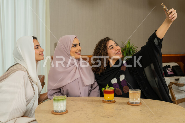 Developing social relationships, enjoying spending time with friends, a fun and acquaintance session on a holiday, taking souvenir selfies, Arab Gulf Emirati friends sitting at a table in a cafe, Saudi women taking a selfie with a mobile phone, a high-end coffee shop in the Emirates