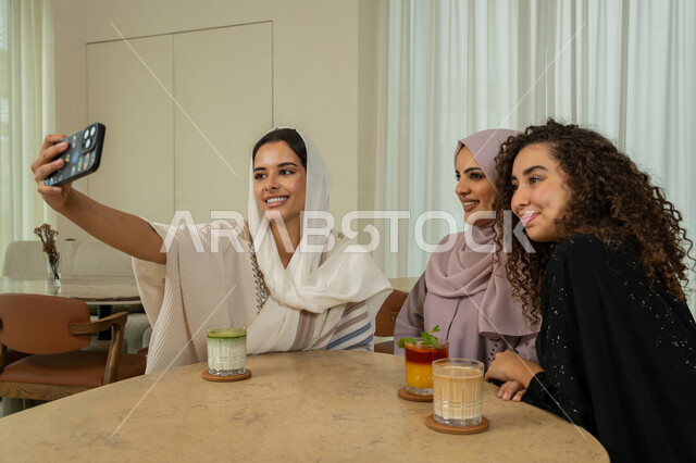 قضاء وقت ممتع برفقة الأصدقاء، التقاط صور السيلفي التذكارية، تطوير العلاقات الإجتماعية، جلسة وناسة وتعارف يوم الاجازة، صديقات عربيات خليجيات اماراتيات يجلسن على طاولة في المقهى، نساء سعوديات يلتقطون صورة الشخصية عبر الهاتف المحمول، كوفي شوب راقي بالامارات