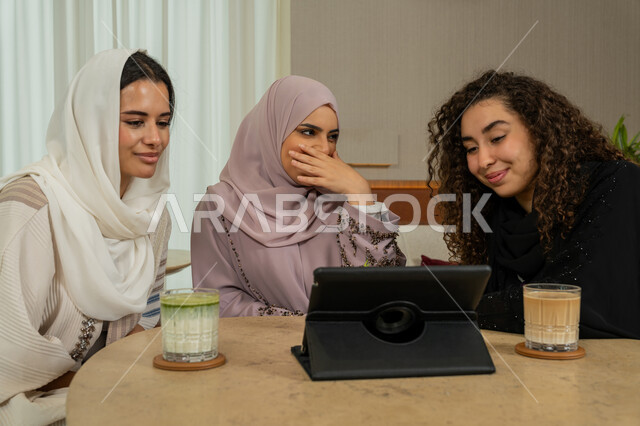 قضاء وقت ممتع مع الأصدقاء، تصفح الإنترنت من جهاز التابلت، ايماءات الدهشة والصدمة، تطوير العلاقات الإجتماعية، صديقات عربيات خليجيات اماراتيات يجلسن على طاولة في المقهى، نساء سعوديات يقومون ببث مباشر عبر مواقع التواصل الاجتماعي، الاستمتاع بمشروب بارد في كوفي شوب راقي
