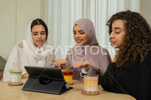 A fun and friendly session, browsing the internet on a tablet, spending quality time with friends, developing social relationships, Arab Gulf Emirati female friends sitting at a table in a cafe, Saudi women broadcasting live on social media, enjoying a cold drink in a high-end coffee shop