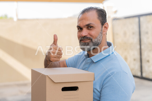 علامة الموافقة و الامتياز, خدمات الشحن والتوصيل ذات الجودة العالية، طلب و تسوق المنتجات عبر الإنترنت، صورة مقربة لمندوب عربي خليجي اماراتي مبتسم يرتدي الزي الكاجوال يحمل صندوق كرتوني، رجل سعودي يقوم برفع اصبع الابهام للاعلى بتعابير الرضا والقبول