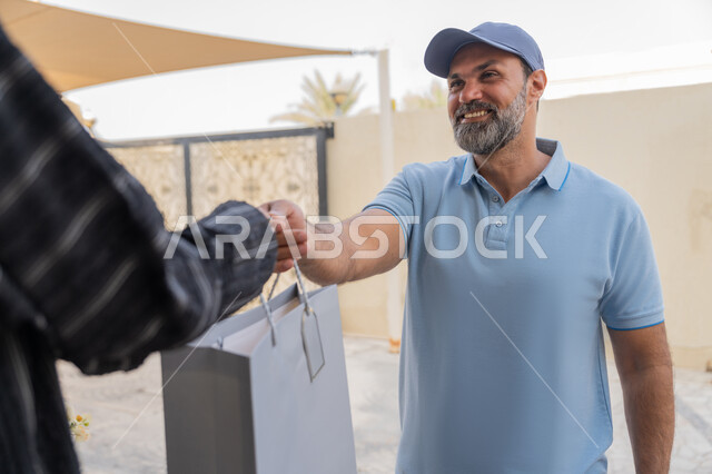 طلب المنتجات والتسوق عبر الإنترنت, شبكة التوصيل الاونلاين في الخليج، خدمة الشحن ذات الجودة العالية، سيدة سعودية تقوم بإستلام البضائع بالوقت المحدد و بشكل سليم، صورة مقربة لامرأة عربية خليجية اماراتية تأخذ كيس ورقي من مندوب عربي خليجي اماراتي يرتدي الزي الكاجوال
