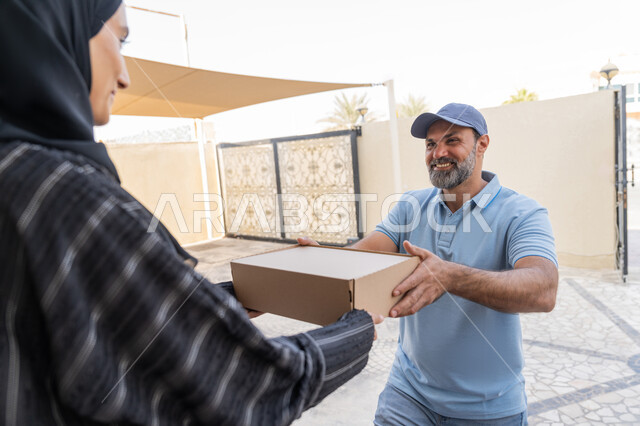 التسوق وطلب المنتجات عبر الإنترنت, شبكة التوصيل الاونلاين في الخليج، خدمة الشحن ذات الجودة العالية، سيدة سعودية تقوم بإستلام البضائع بالوقت المحدد و بشكل سليم، صورة مقربة لامرأة عربية خليجية اماراتية تأخذ صندوق كرتوني من مندوب عربي خليجي اماراتي يرتدي الزي الكاجوال