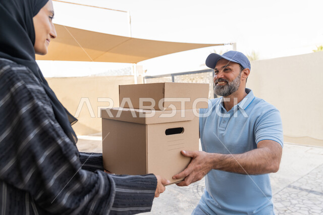 شبكة التوصيل الاونلاين في الخليج، خدمة الشحن ذات الجودة العالية، طلب المنتجات والتسوق عبر الإنترنت, سيدة سعودية تقوم بإستلام البضائع بالوقت المحدد و بشكل سليم، صورة مقربة لامرأة عربية خليجية اماراتية تأخذ صناديق كرتونية من مندوب عربي خليجي اماراتي يرتدي الزي الكاجوال