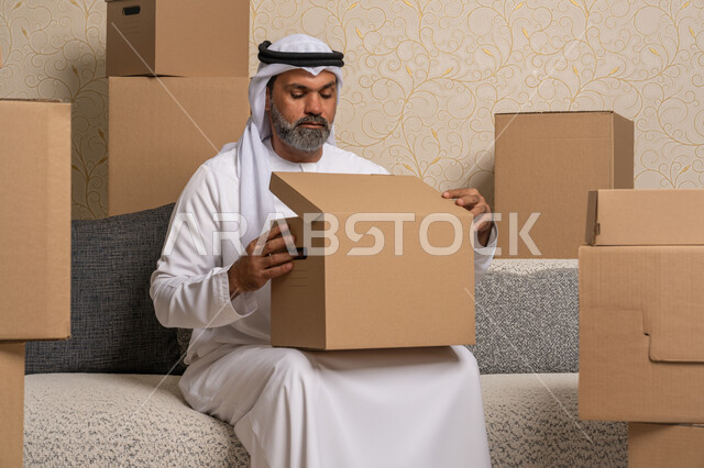 ترتيب وتنظيم المنزل، نقل صناديق الأغراض المنزلية، ايماءات السعادة والسرور لامتلاك سكن جديد، خدمات النقل والتوصيل السريع، مفهوم الاسكان والتمليك والاستقرار، رجل عربي خليجي اماراتي يرتدي الكندورة والغترة يجلس على اريكة في البيت الجديد ويقوم بفتح صندوق كرتوني
