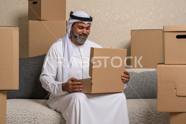 نقل صناديق الأغراض المنزلية، ايماءات السعادة والسرور لامتلاك سكن جديد، خدمات النقل والتوصيل السريع، مفهوم الاسكان والتمليك والاستقرار، ترتيب وتنظيم المنزل، رجل عربي خليجي اماراتي يرتدي الكندورة والغترة يجلس على اريكة في البيت الجديد ويقوم بفتح صندوق كرتوني