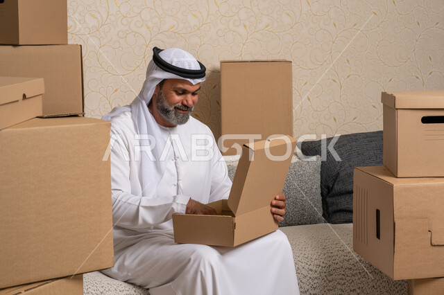 مفهوم الاسكان والتمليك والاستقرار، ترتيب وتنظيم المنزل، نقل صناديق الأغراض المنزلية، تعابير السعادة والسرور لامتلاك سكن جديد، خدمات النقل والتوصيل السريع، رجل عربي خليجي اماراتي يرتدي الكندورة والغترة يجلس على اريكة في البيت الجديد ويقوم بفتح صندوق كرتوني