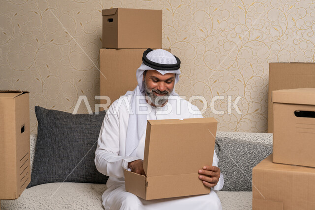 ترتيب وتنظيم المنزل، نقل صناديق الأغراض المنزلية، تعابير السعادة والسرور لامتلاك سكن جديد، خدمات النقل والتوصيل السريع، مفهوم الاسكان والتمليك والاستقرار، رجل عربي خليجي اماراتي يرتدي الكندورة والغترة يجلس على اريكة في البيت الجديد ويقوم بفتح صندوق كرتوني