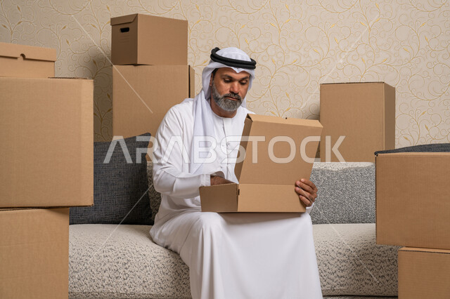تنظيم وترتيب المنزل، نقل صناديق الأغراض المنزلية، تعابير السعادة والسرور لامتلاك سكن جديد، خدمات النقل والتوصيل السريع، مفهوم الاسكان والتمليك والاستقرار، رجل عربي خليجي اماراتي يرتدي الكندورة والغترة يجلس على اريكة في البيت الجديد ويقوم بفتح صندوق كرتوني