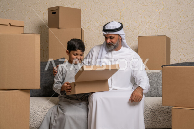 نقل صناديق الأغراض المنزلية، مفهوم الاسكان والتمليك والاستقرارالعائلي، ترتيب وتنظيم المنزل، امتلاك سكن جديد، رجل عربي خليجي اماراتي يرتدي الكندورة والغترة يجلس على اريكة في البيت الجديد يقوم بفتح صندوق كرتوني مع ابنه