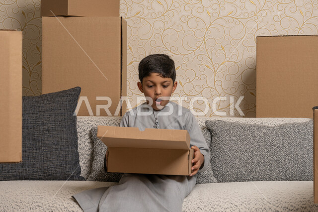 خدمات النقل والتوصيل السريع، مفهوم الاسكان والتمليك والاستقرار، ترتيب وتنظيم المنزل، نقل صناديق الأغراض المنزلية، امتلاك سكن جديد، ولد عربي خليجي اماراتي يرتدي الكندورة الرمادية يجلس على اريكة في البيت الجديد يقوم بفتح صندوق كرتوني بايماءات الدهشة