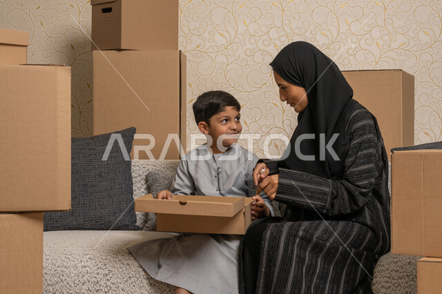 ترتيب وتنظيم المنزل، تعابير السعادة والسرور لامتلاك سكن جديد، مفهوم الاسكان والتمليك والاستقرار العائلي، نقل صناديق الأغراض المنزلية، امرأة عربية خليجية اماراتية تجلس على اريكة في البيت الجديد مع ابنها ويقومان بفتح صندوق كرتوني
