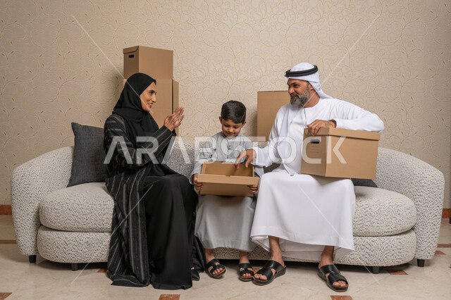 مفهوم الاسكان والتمليك والاستقرار العائلي، نقل صناديق الأغراض المنزلية، ترتيب وتنظيم المنزل، ايماءات السعادة لامتلاك سكن جديد، عائلة عربية خليجية اماراتية يجلسون على اريكة في البيت الجديد وبجانبهما حاويات كرتونية، الوحدة العائلية والترابط الأسري