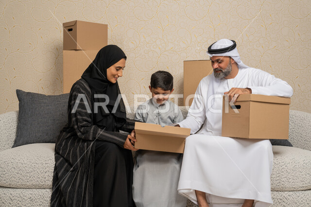 Moving boxes of household items, arranging and organizing the house, gestures of happiness for owning a new home, an Arab Gulf Emirati family sitting on a sofa in the new house with cardboard containers next to them, the concept of housing, ownership and family stability, family unity and family cohesion