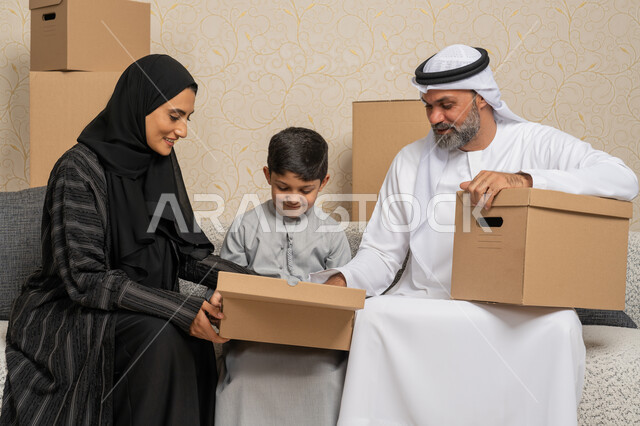 نقل صناديق الأغراض المنزلية، ترتيب وتنظيم المنزل، ايماءات السعادة لامتلاك سكن جديد، عائلة عربية خليجية اماراتية يجلسون على اريكة في البيت الجديد وبجانبهم حاويات كرتونية، مفهوم الاسكان والتمليك والاستقرار العائلي، الوحدة العائلية والترابط الأسري