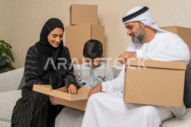 Arranging and organizing the house, gestures of happiness for owning a new home, moving boxes of household items, an Arab Gulf Emirati family sitting on a sofa in the new house with cardboard containers next to them, the concept of housing, ownership and family stability, family unity and family cohesion