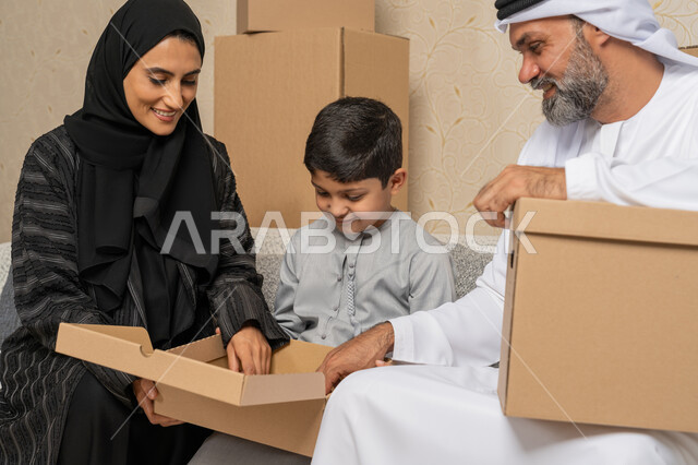 تنظيم وترتيب المنزل، ايماءات السعادة لامتلاك سكن جديد، نقل صناديق الأغراض المنزلية، عائلة عربية خليجية اماراتية يجلسون على اريكة في البيت الجديد وبجانبهم حاويات كرتونية، مفهوم الاسكان والتمليك والاستقرار العائلي، الوحدة العائلية والترابط الأسري