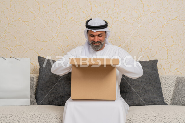 طلب المنتجات والتسوق عبر الإنترنت، استلام الطلبات الاونلاين في المنزل، عملية توصيل الطلبات للزبائن، الشحن والتوصيل ذات الجودة العالية، رجل عربي خليجي اماراتي يرتدي الكندورة والغترة مبتسم يجلس على اريكة في فرفة المعيشة ويقوم بفتح صندوق كرتوني