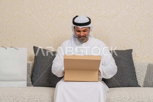 Shopping and ordering products online, receiving online orders at home, customer delivery process, high quality shipping and delivery, an Arab Gulf Emirati man wearing a kandura and a ghutra smiling sitting on a sofa in the living room and opening a cardboard box