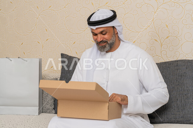 استلام الطلبات الاونلاين في المنزل، عملية توصيل الطلبات للزبائن، الشحن والتوصيل ذات الجودة العالية، التسوق وطلب المنتجات عبر الإنترنت، رجل عربي خليجي اماراتي يرتدي الكندورة والغترة مبتسم يجلس على اريكة في فرفة المعيشة ويقوم بفتح صندوق كرتوني