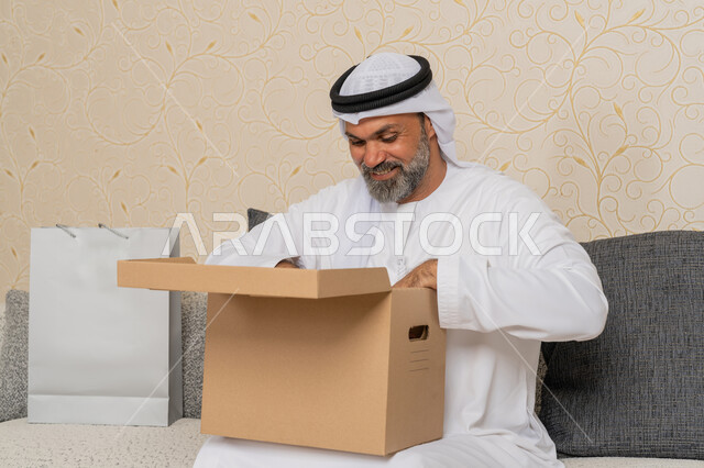 عملية توصيل الطلبات للزبائن، الشحن والتوصيل ذات الجودة العالية، التسوق وطلب المنتجات عبر الإنترنت، رجل عربي خليجي اماراتي يرتدي الكندورة والغترة مبتسم يجلس على اريكة في فرفة المعيشة ويقوم بفتح صندوق كرتوني، استلام الطلبات الاونلاين في المنزل