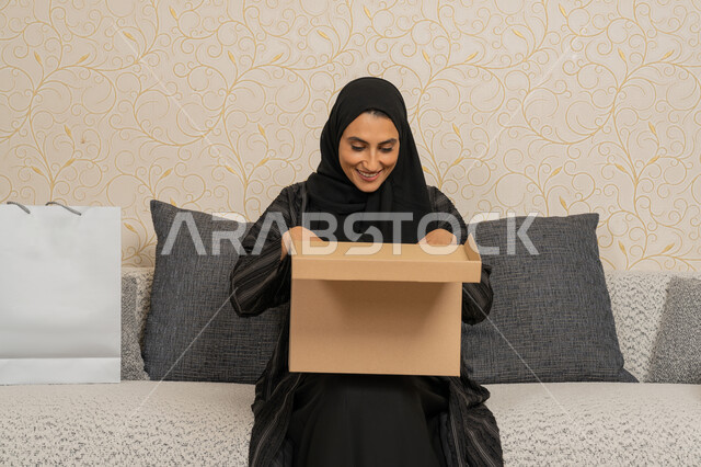 الشحن والتوصيل ذات الجودة العالية، التسوق وطلب المنتجات عبر الإنترنت، عملية توصيل الطلبات للزبائن، سيده عربيه خليجيه اماراتية محجبه مبتسمه تجلس على اريكة في فرفة المعيشة، امرأة سعودية تحمل بيدها صندوق كرتوني، استلام الطلبات الاونلاين في المنزل