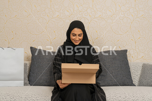 High quality delivery and shipping, online shopping and ordering, customer delivery process, smiling veiled Emirati Gulf Arab lady sitting on a sofa in living room, Saudi woman holding a cardboard box, receiving online orders at home