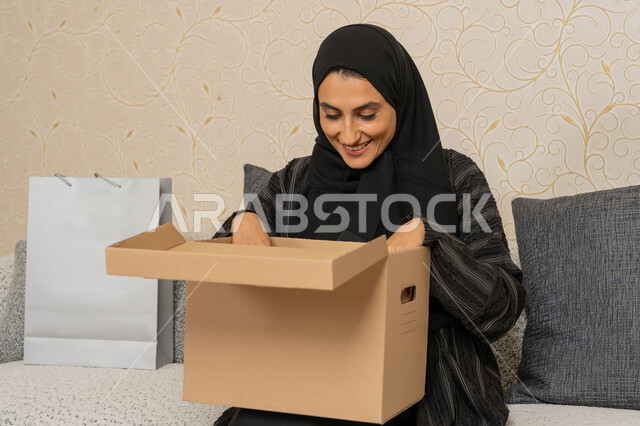 طلب المنتجات والتسوق عبر الإنترنت، التوصيل والشحن ذات الجودة العالية، عملية توصيل الطلبات للزبائن، سيده عربيه خليجيه اماراتية محجبه مبتسمه تجلس على اريكة في فرفة المعيشة، امرأة سعودية تحمل بيدها صندوق كرتوني، استلام الطلبات الاونلاين في المنزل