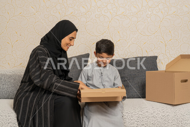 مفهوم الاسكان والتمليك والاستقرار العائلي، ترتيب وتنظيم المنزل، التشارك والتعاون في نقل صناديق الأغراض المنزلية، ايماءات السعادة والسرور لامتلاك سكن جديد، امرأة عربية خليجية اماراتية تجلس على اريكة في البيت الجديد مع ابنها ويقومان بفتح صندوق كرتوني