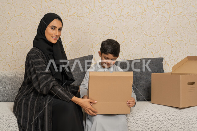 Organizing and arranging the house, sharing and cooperating in moving boxes of household items, gestures of happiness and joy for owning a new home, an Emirati Gulf Arab woman sitting on a sofa in the new house with her son and they open a cardboard box, the concept of housing, ownership and family stability