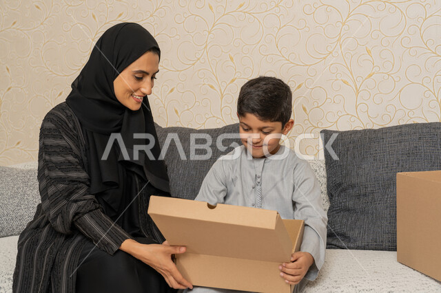 Organizing and arranging the house, sharing and cooperating in moving boxes of household items, gestures of happiness and joy for owning a new home, an Arab Gulf Emirati woman sitting on a sofa in the new house with her son and they open a cardboard box, the concept of housing, ownership and family stability