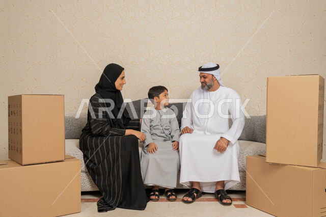 تعابير السعادة لامتلاك سكن جديد، تنظيم وترتيب المنزل، نقل صناديق الأغراض المنزلية، عائلة عربية خليجية اماراتية يجلسون على اريكة في البيت الجديد وبجانبهم حاويات كرتونية، مفهوم الاسكان والتمليك والاستقرار العائلي، الوحدة العائلية والترابط الأسري