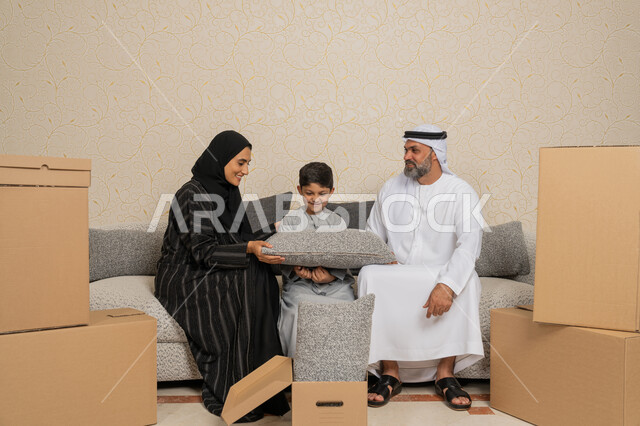 ايماءات السعادة لامتلاك سكن جديد، تنظيم وترتيب المنزل، نقل صناديق الاغراض المنزلية، عائلة عربية خليجية اماراتية يجلسون على اريكة في البيت الجديد وبجانبهم حاويات كرتونية، مفهوم الاسكان والتمليك والاستقرار العائلي، الوحدة العائلية والترابط الأسري