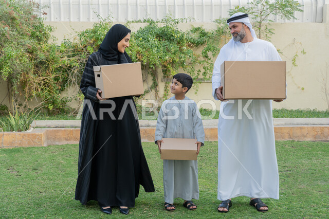 ايماءات السعادة والسرور بامتلاك سكن جديد، التعاون والتشارك في نقل صناديق الأغراض المنزلية، عائلة عربية خليجية اماراتية يقفون في الحديقة الخارجية للبيت الجديد ويحملون حاويات كرتونية، ترتيب وتنظيم المنزل، مفهوم الاسكان والتمليك والاستقرار العائلي