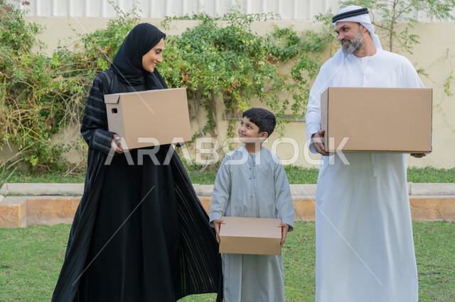 التعاون والتشارك في نقل صناديق الأغراض المنزلية، عائلة عربية خليجية اماراتية يقفون في الحديقة الخارجية للبيت الجديد ويحملون حاويات كرتونية، ايماءات السعادة والسرور بامتلاك سكن جديد، ترتيب وتنظيم المنزل، مفهوم الاسكان والتمليك والاستقرار العائلي