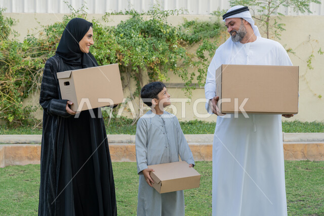 التشارك والتعاون في نقل صناديق الأغراض المنزلية، عائلة عربية خليجية اماراتية يقفون في الحديقة الخارجية للبيت الجديد ويحملون حاويات كرتونية، ايماءات السعادة والسرور بامتلاك سكن جديد، ترتيب وتنظيم المنزل، مفهوم الاسكان والتمليك والاستقرار العائلي