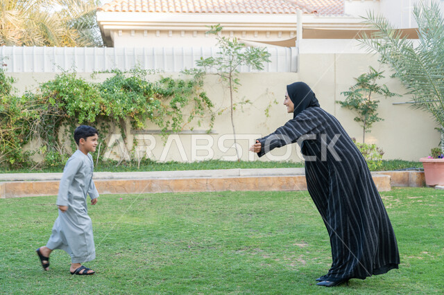 قضاء وقت ممتع ومسلي في المنتزه، جو عائلي مليء بالحب، التنزه في الهواء الطلق في الحديقة, الاعتناء بالاطفال والاهتمام بهم، امرأة عربية خليجية اماراتية محجبة ترتدي عباءة سوداء تفتح يديها لاستقبال ابنها بالحضن، الألفة والمحبة والترابط الأسري، تعابير السعادة والسرور