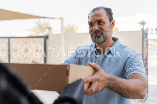 خدمة الشحن ذات الجودة العالية، شبكة التوصيل الاونلاين في الخليج، التسوق وطلب المنتجات عبر الإنترنت, مندوب عربي خليجي اماراتي يرتدي الزي الكاجوال يقوم باعطاء صناديق كرتونية لامرأة عربية خليجية امارتيه، سيدة سعودية تقوم بإستلام البضائع بالوقت المحدد و بشكل سليم
