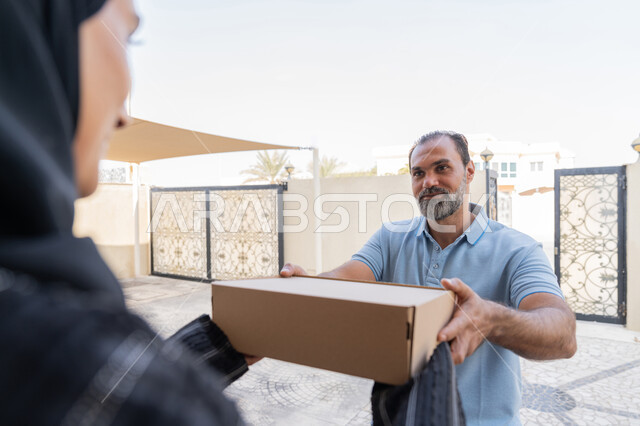 شبكة التوصيل الاونلاين في الخليج، التسوق وطلب المنتجات عبر الإنترنت, خدمة الشحن ذات الجودة العالية، مندوب عربي خليجي اماراتي يرتدي الزي الكاجوال يقوم باعطاء صناديق كرتونية لامرأة عربية خليجية امارتيه، سيدة سعودية تقوم بإستلام البضائع بالوقت المحدد و بشكل سليم
