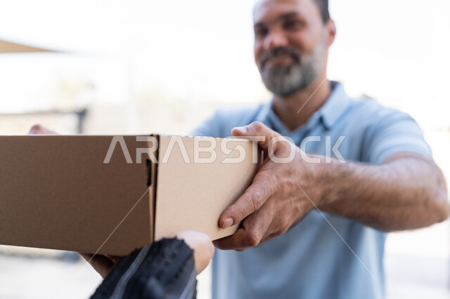 التسوق وطلب المنتجات عبر الإنترنت, خدمة الشحن ذات الجودة العالية، شبكة التوصيل الاونلاين في الخليج، مندوب عربي خليجي اماراتي يرتدي الزي الكاجوال يقوم باعطاء صناديق كرتونية لامرأة عربية خليجية امارتيه، سيدة سعودية تقوم بإستلام البضائع بالوقت المحدد و بشكل سليم