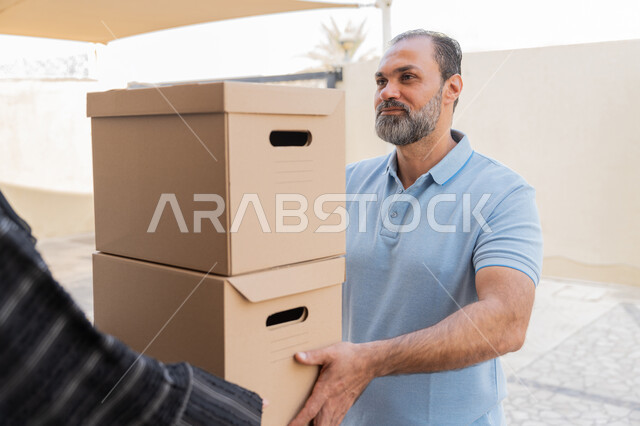 طلب المنتجات والتسوق عبر الإنترنت, شبكة التوصيل الاونلاين في الخليج، مندوب عربي خليجي اماراتي يرتدي الزي الكاجوال يقوم باعطاء صناديق كرتونية لامرأة عربية خليجية امارتيه، خدمة الشحن ذات الجودة العالية، سيدة سعودية تقوم بإستلام البضائع بالوقت المحدد و بشكل سليم
