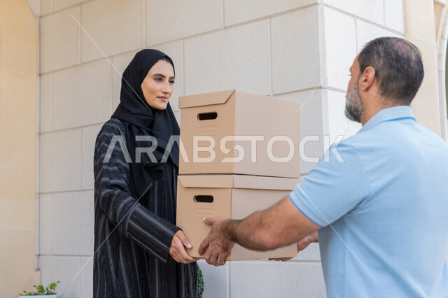 التسوق وطلب المنتجات عبر الإنترنت, شبكة التوصيل الاونلاين في الخليج، مندوب عربي خليجي اماراتي يرتدي الزي الكاجوال يقوم باعطاء صناديق كرتونية لامرأة عربية خليجية امارتيه، خدمة الشحن ذات الجودة العالية، سيدة سعودية تقوم بإستلام البضائع بالوقت المحدد و بشكل سليم