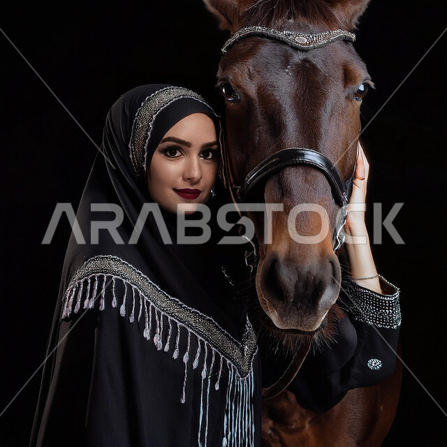 Equestrianism and horse riding hobby, fun recreational activity, interest and passion for horse riding, Saudi Arabian people's attachment to horses, close-up portrait of a Saudi Arabian Gulf girl wearing a black abaya and standing next to a purebred Arabian horse, black background