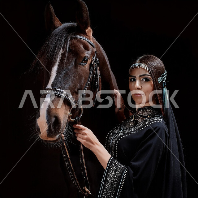 الفروسية وهواية ركوب الأحصنة، نشاط ترفيهي ممتع، الاهتمام والشغف بركوب الجياد، تعلق أبناء المملكة العربية السعودية بالخيول، بورتريه مقرب لفتاة عربية سعودية خليجية ترتدي العباءة السوداء و تقف بجانب خيل عربي اصيل، خلفية سوداء