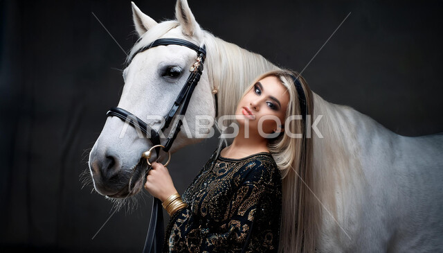 نشاط ترفيهي ممتع، الفروسية وهواية ركوب الأحصنة، الاهتمام والشغف بركوب الجياد، تعلق أبناء المملكة العربية السعودية بالخيول، فتاة عربية سعودية خليجية ترتدي العباءة السوداء و تقف بجانب خيل عربي اصيل، خلفية رمادية