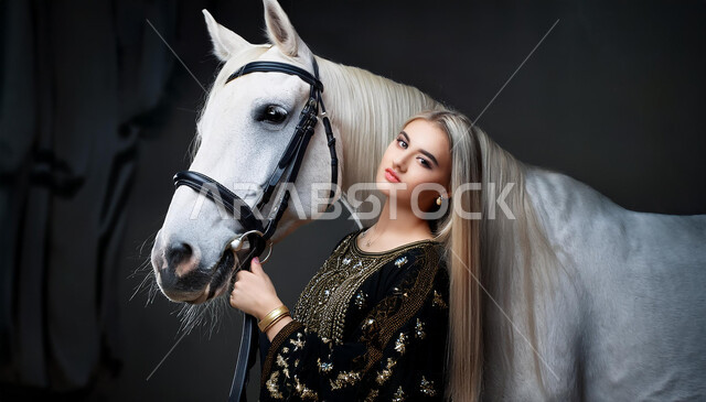 نشاط ترفيهي ممتع، الفروسية وهواية ركوب الأحصنة، الاهتمام والشغف بركوب الجياد، تعلق أبناء المملكة العربية السعودية بالخيول، فتاة عربية سعودية خليجية ترتدي العباءة السوداء و تقف بجانب خيل عربي اصيل، خلفية رمادية