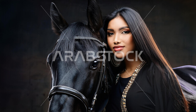 نشاط ترفيهي ممتع، الفروسية وهواية ركوب الأحصنة، الاهتمام والشغف بركوب الجياد، تعلق أبناء المملكة العربية السعودية بالخيول، فتاة عربية سعودية خليجية ترتدي العباءة السوداء و تقف بجانب خيل عربي اصيل، خلفية رمادية
