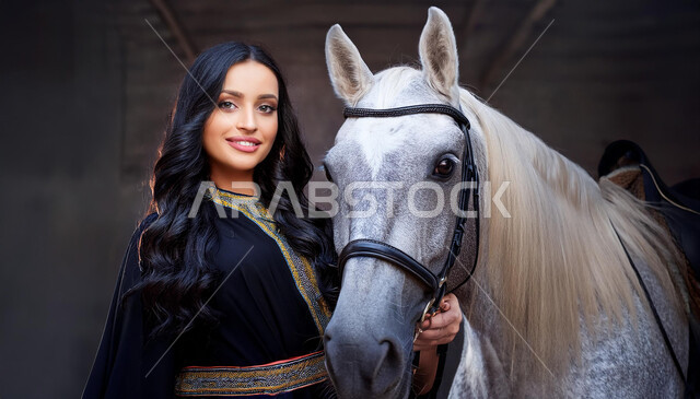 Fun recreational activity, equestrianism and horse riding hobby, interest and passion for horse riding, Saudi Arabian people's attachment to horses, Saudi Arabian Gulf girl wearing black abaya and standing next to purebred Arabian horse, gray background