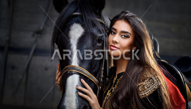 نشاط ترفيهي ممتع، الفروسية وهواية ركوب الأحصنة، الاهتمام والشغف بركوب الجياد، تعلق أبناء المملكة العربية السعودية بالخيول، فتاة عربية سعودية خليجية ترتدي العباءة السوداء و تقف بجانب خيل عربي اصيل، خلفية رمادية