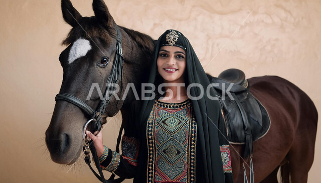 تعلق أبناء المملكة العربية السعودية بالخيول، نشاط ترفيهي ممتع، الفروسية وهواية ركوب الأحصنة، الاهتمام والشغف بركوب الجياد، فتاة عربية سعودية خليجية ترتدي العباءة السوداء و تقف بجانب خيل عربي اصيل